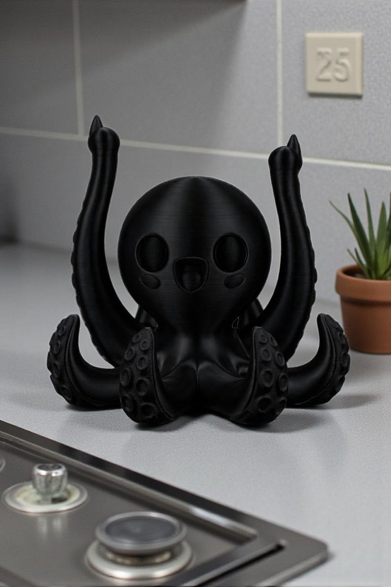 Puede incluir: Figura de pulpo negro, impresa en 3D, con una cara sonriente y tent&aacute;culos levantados. La figura est&aacute; sobre una superficie clara, con una peque&ntilde;a planta en maceta y el n&uacute;mero 25 en la pared. El pulpo tiene un dise&ntilde;o lindo y de dibujos animados.