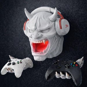 Può includere: Una testa di demone bianca con corna, una bocca rossa incandescente e cuffie, insieme a due supporti per controller di gioco. Un supporto è bianco, l'altro nero. Le cuffie hanno dettagli rossi e il marchio "HyperX".