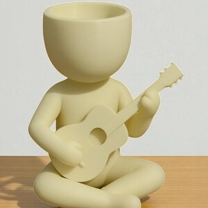 May include: A light yellow figurine of a person seated cross-legged, playing a guitar. The figurine has a large head with a hollowed-out top, possibly for use as a planter. The guitar is also light yellow, with a round sound hole.