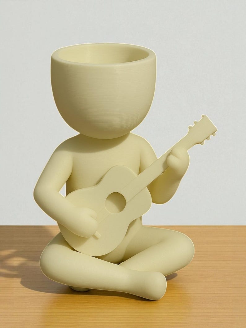 May include: A light yellow figurine of a person seated cross-legged, playing a guitar. The figurine has a large head with a hollowed-out top, possibly for use as a planter. The guitar is also light yellow, with a round sound hole.