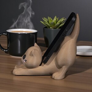 May include: A tan cat-shaped phone holder with a black phone in place. The cat is in a playful pose, with its tail up. A black mug with coffee and steam, a small potted succulent, and a white plate are in the background.