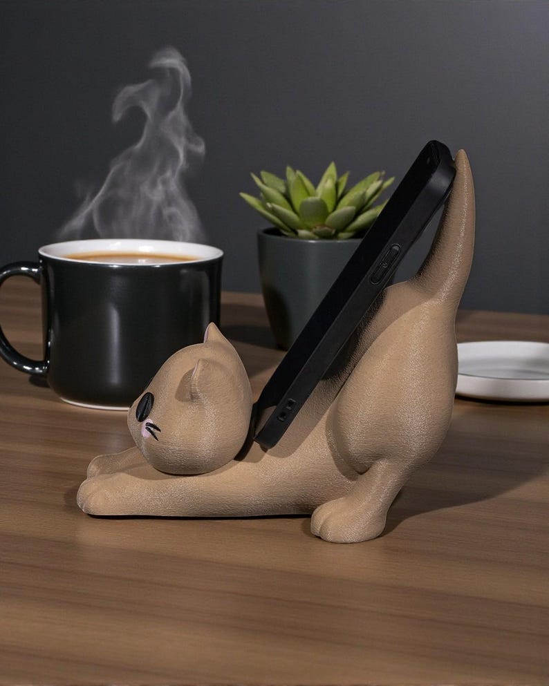 May include: A tan cat-shaped phone holder with a black phone in place. The cat is in a playful pose, with its tail up. A black mug with coffee and steam, a small potted succulent, and a white plate are in the background.