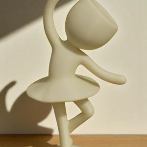 May include: A cream-colored figurine of a ballerina in a graceful pose. The figure has a rounded head, outstretched arms, and a tutu. The ballerina is balanced on one leg, with the other bent. The figurine is on a round base.
