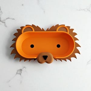 May include: A novelty lion-shaped tray in orange and brown. The tray features a brown mane, an orange central section, and a lion's face with black eyes and a brown nose. Ideal for holding small items, it measures approximately 25 cm in length.