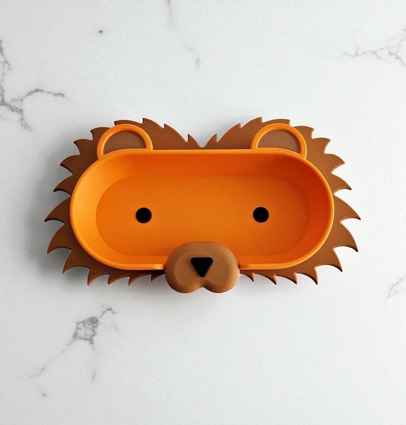 May include: A novelty lion-shaped tray in orange and brown. The tray features a brown mane, an orange central section, and a lion's face with black eyes and a brown nose. Ideal for holding small items, it measures approximately 25 cm in length.