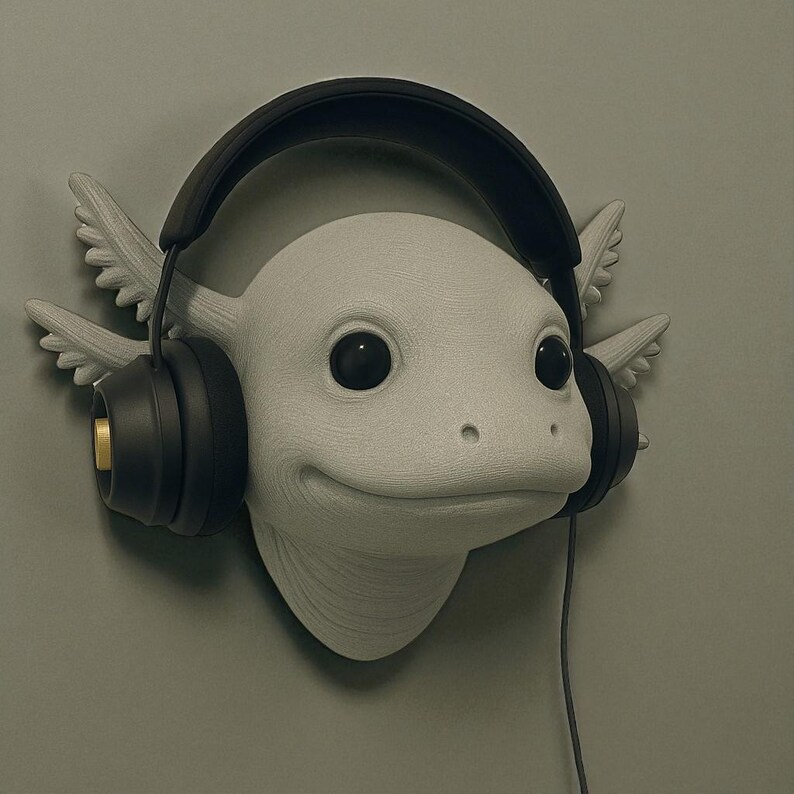 Puede incluir: Escultura de cabeza de ajolote blanco con grandes ojos negros y expresi&oacute;n sonriente, con auriculares negros. El ajolote tiene branquias plumosas y est&aacute; montado en una pared verde apagada. Los auriculares tienen detalles dorados.