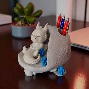K&ouml;nnte beinhalten: Ein wei&szlig;er Drachen-Figur-Stifthalter mit einem schlafenden Drachen und blauen Kristallakzenten. Der Stifthalter hat ein graues, steinartiges Aussehen und h&auml;lt rote und blaue Stifte. Der Drache befindet sich in einer entspannten Pose.