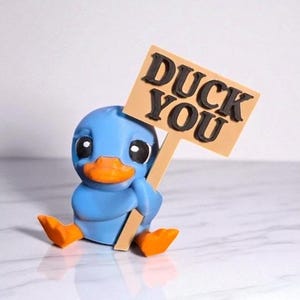 May include: A blue duck figurine with an orange beak and feet, holding a sign that reads "DUCK YOU" in black letters. The duck is sitting down, and the sign is tan with a brown border.
