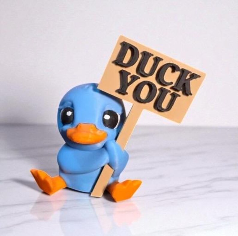 May include: A blue duck figurine with an orange beak and feet, holding a sign that reads "DUCK YOU" in black letters. The duck is sitting down, and the sign is tan with a brown border.