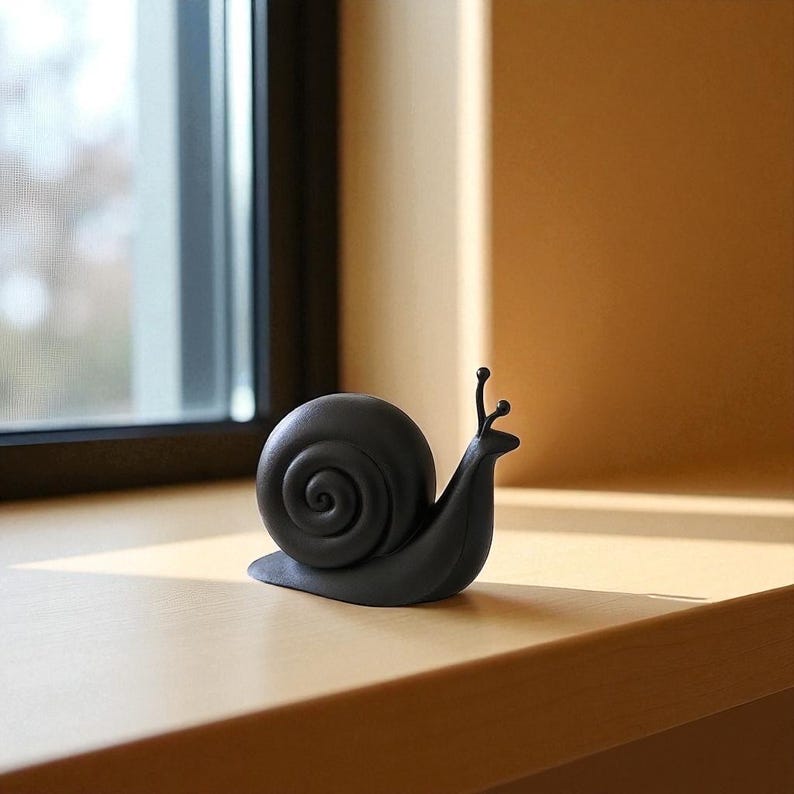 May include: A black snail ornament with a coiled shell and two small antennae. The snail is placed on a wooden windowsill, with a window and natural light in the background. The figurine is a decorative item.