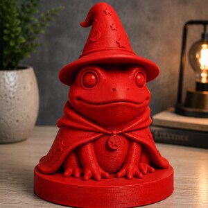 May include: A red frog figurine wearing a wizard hat and cape. The hat features crescent moon and star details. The frog is on a round base and has a star pendant on its cape. The figurine is a whimsical decorative item.