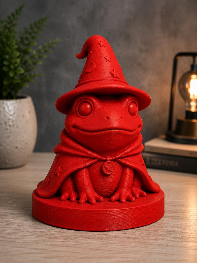 May include: A red frog figurine wearing a wizard hat and cape. The hat features crescent moon and star details. The frog is on a round base and has a star pendant on its cape. The figurine is a whimsical decorative item.