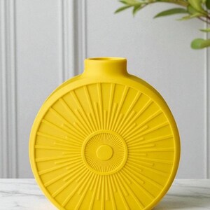 May include: A bright yellow, round vase with a sunburst design. The vase has a narrow neck and a flat base. The sunburst pattern radiates from a central circle, creating a textured surface. The vase is made of ceramic or a similar material.