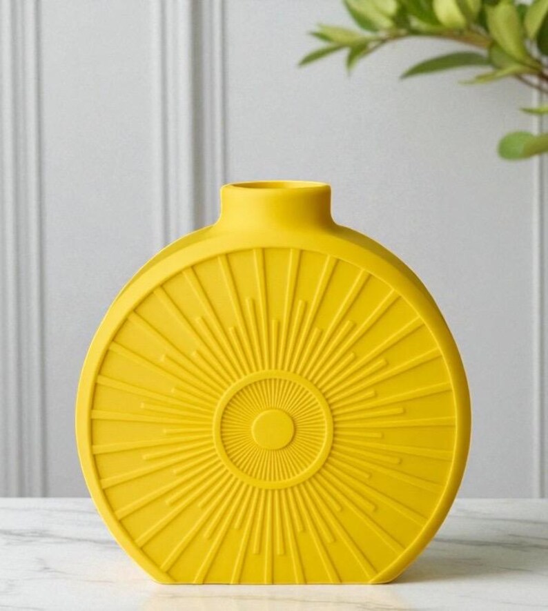May include: A bright yellow, round vase with a sunburst design. The vase has a narrow neck and a flat base. The sunburst pattern radiates from a central circle, creating a textured surface. The vase is made of ceramic or a similar material.