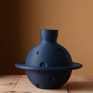 Könnte beinhalten: Eine dunkelblaue Keramikvase mit einem planetenähnlichen Design. Die Vase hat einen runden Sockel mit einem Ring darum, einen kugelförmigen Körper mit mehreren kreisförmigen Vertiefungen und einen zylindrischen Hals. Die Vase steht auf einer Holzoberfläche vor einem braunen Hintergrund.
