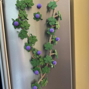 May include: A decorative vine with green leaves and purple berries, attached to a stainless steel surface. The vine is made of plastic and is likely used for home decor or crafts. The image is well-lit and the colors are vibrant.
