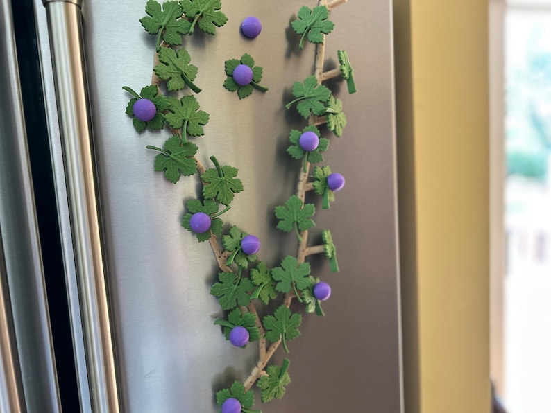 May include: A decorative vine with green leaves and purple berries, attached to a stainless steel surface. The vine is made of plastic and is likely used for home decor or crafts. The image is well-lit and the colors are vibrant.