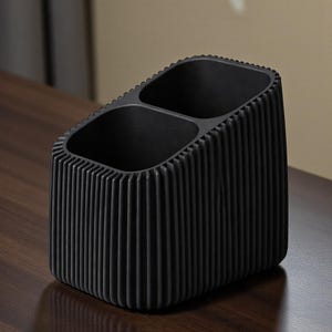 May include: A black desk organizer with two compartments. The organizer has a rectangular shape with vertical ribbed detailing on the sides. The interior is smooth and the exterior has a textured, ridged design.