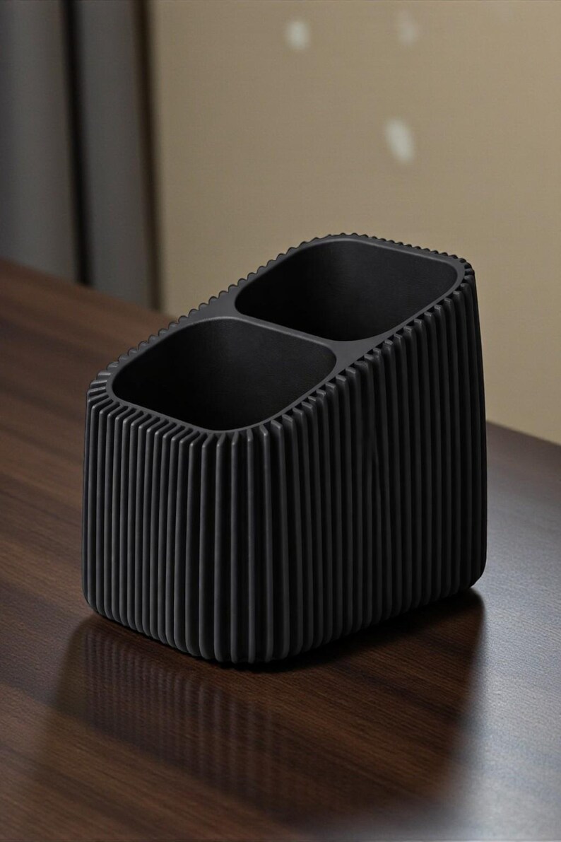 May include: A black desk organizer with two compartments. The organizer has a rectangular shape with vertical ribbed detailing on the sides. The interior is smooth and the exterior has a textured, ridged design.