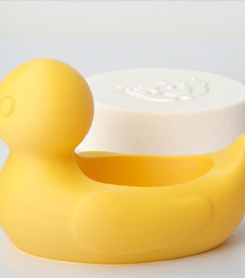 Op de afbeelding: Een gele rubberen eendvormige zeepbakje met een witte, ronde zeep in de achtergrond. De eend heeft een uitgesneden midden om zeep in te bewaren. De zeep heeft een reli&euml;f ontwerp.