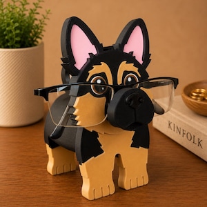 Pode incluir: Um suporte de &oacute;culos de madeira em forma de c&atilde;o pastor alem&atilde;o. O c&atilde;o &eacute; preto, castanho claro e rosa, com um par de &oacute;culos apoiados no nariz. O suporte est&aacute; sobre uma superf&iacute;cie de madeira, com uma planta e um livro ao fundo.