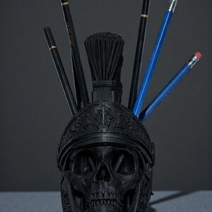 Puede incluir: Un organizador de escritorio negro con forma de calavera con un dise&ntilde;o de casco romano. El organizador contiene l&aacute;pices y un pincel. Los l&aacute;pices son negros y azules con detalles dorados y gomas rojas. El accesorio de escritorio est&aacute; sobre una superficie gris.