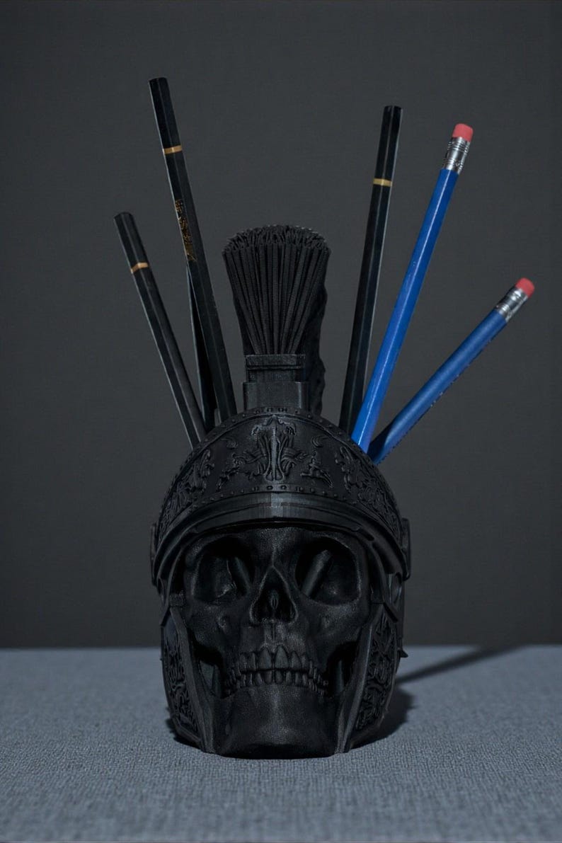 Puede incluir: Un organizador de escritorio negro con forma de calavera con un dise&ntilde;o de casco romano. El organizador contiene l&aacute;pices y un pincel. Los l&aacute;pices son negros y azules con detalles dorados y gomas rojas. El accesorio de escritorio est&aacute; sobre una superficie gris.