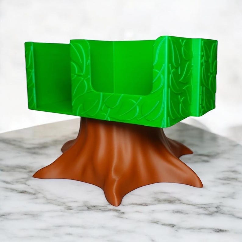 May include: A decorative desk organizer shaped like a tree. The base is brown, resembling a tree trunk, and supports a bright green, leaf-patterned organizer with two compartments. The organizer is designed to hold small items.