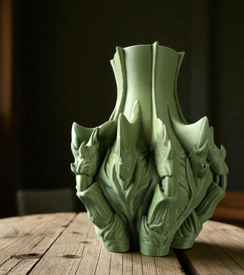 May include: A green ceramic vase with a unique, textured design. The vase has a flared top and a base with multiple points. The surface features intricate, leaf-like patterns, creating a sculptural appearance. The vase is displayed on a wooden surface.
