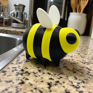 May include: A 3D-printed bee figurine with a yellow and black striped body, black legs, and white wings. The bee has a rounded shape and a large, circular eye. This decorative item is suitable for home decor.