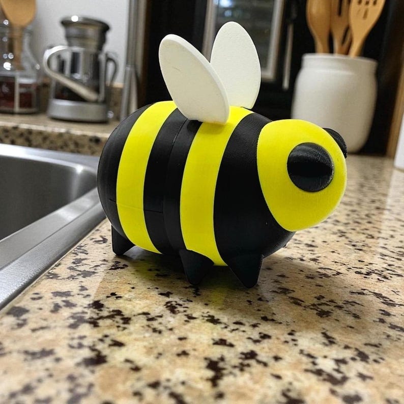 May include: A 3D-printed bee figurine with a yellow and black striped body, black legs, and white wings. The bee has a rounded shape and a large, circular eye. This decorative item is suitable for home decor.