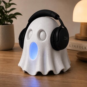 May include: A white ghost-shaped speaker with a blue glowing mouth, wearing black headphones. The speaker has two round eye-like features. The headphones are black with the brand name "HyperX" visible on the earcups. The speaker is on a wooden surface.