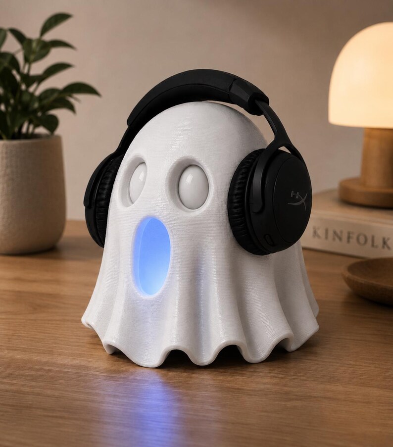 May include: A white ghost-shaped speaker with a blue glowing mouth, wearing black headphones. The speaker has two round eye-like features. The headphones are black with the brand name "HyperX" visible on the earcups. The speaker is on a wooden surface.