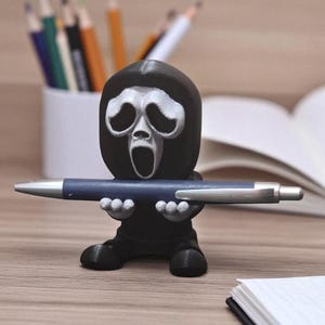 Puede incluir: Un portal&aacute;pices negro con forma de m&aacute;scara de Ghostface de las pel&iacute;culas Scream, sosteniendo un bol&iacute;grafo azul. El portal&aacute;pices tiene una cara blanca y est&aacute; sobre una superficie de madera. Hay l&aacute;pices en una taza blanca en el fondo.
