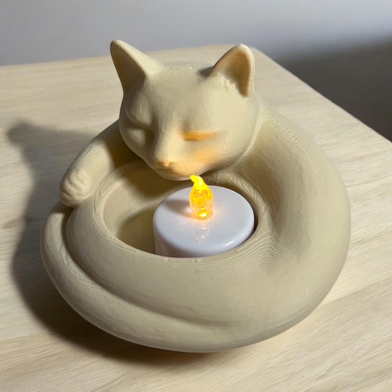 May include: A cream-colored cat-shaped candle holder with a sleeping cat design. The cat's body curls around a white candle with a flickering yellow LED flame. The candle holder is on a light-colored wooden surface.