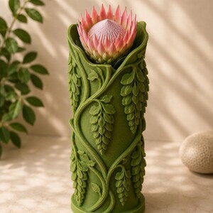 Puede incluir: Un jarr&oacute;n cil&iacute;ndrico verde con un relieve detallado de vides y hojas. Una gran flor de protea rosa y blanca florece desde la parte superior. El jarr&oacute;n mide aproximadamente 20 cm de alto.