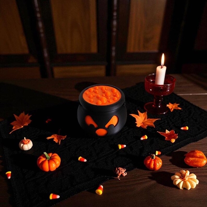 May include: A Halloween-themed still life featuring a black cauldron with an orange interior and jack-o'-lantern face. The scene includes a lit candle in a red glass, decorative pumpkins, fall leaves, and candy corn on a black cloth.