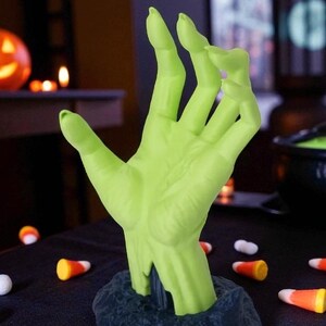 Puede incluir: Una mano verde, similar a la de un zombi, que emerge de una base oscura. La mano es de un verde brillante con una escultura detallada. Caramelos de ma&iacute;z y caramelos blancos est&aacute;n esparcidos sobre la superficie negra, sugiriendo un tema de Halloween.