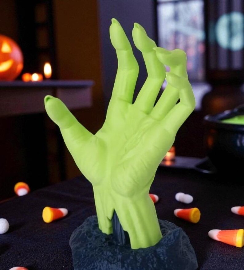 Puede incluir: Una mano verde, similar a la de un zombi, que emerge de una base oscura. La mano es de un verde brillante con una escultura detallada. Caramelos de ma&iacute;z y caramelos blancos est&aacute;n esparcidos sobre la superficie negra, sugiriendo un tema de Halloween.