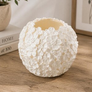 May include: A white, spherical decorative vase covered in small, three-dimensional flower blossoms. The vase has a smooth, off-white interior and sits on a wooden surface. The floral design gives the vase a textured appearance.