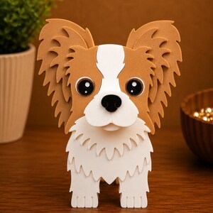 May include: A 3D papercraft dog figurine with large, expressive eyes and oversized, feathery ears. The dog has a white and brown colour scheme, with a white muzzle and chest. The figurine is standing upright on a wooden surface.