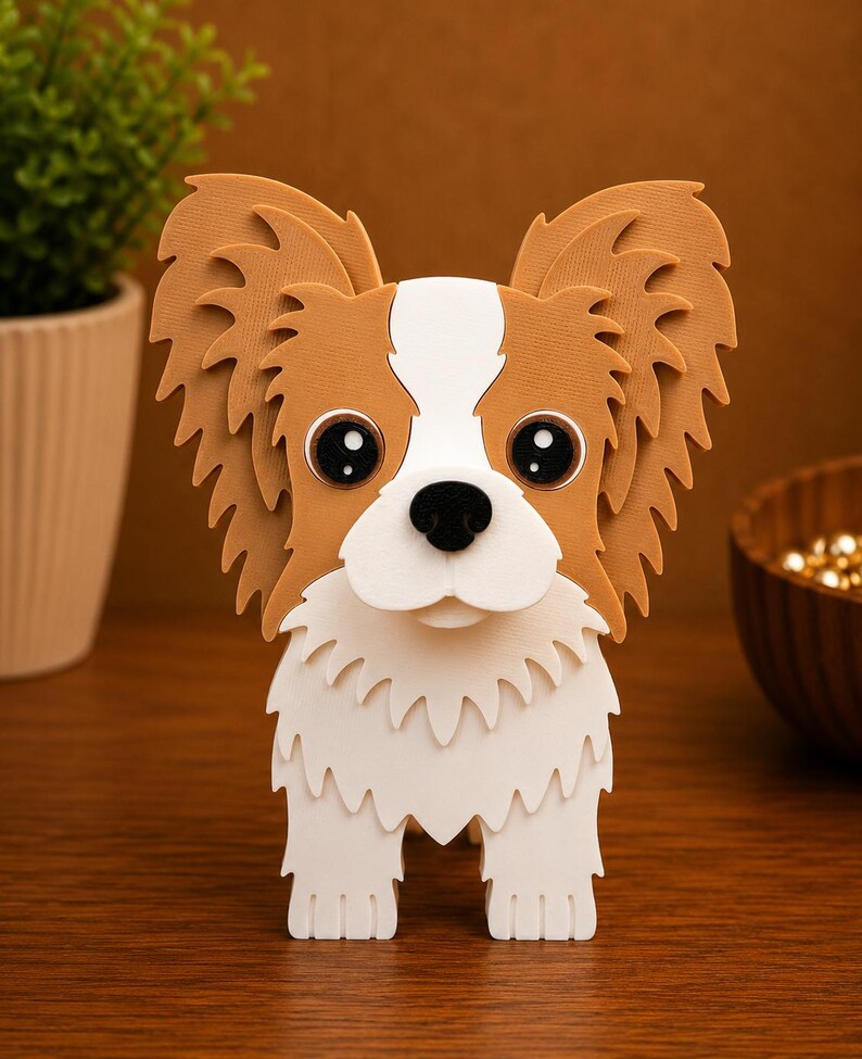 May include: A 3D papercraft dog figurine with large, expressive eyes and oversized, feathery ears. The dog has a white and brown colour scheme, with a white muzzle and chest. The figurine is standing upright on a wooden surface.