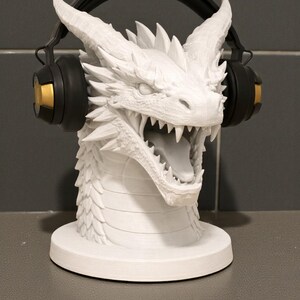 May include: A white dragon head sculpture with detailed scales and sharp teeth, designed to hold headphones. The dragon's head is mounted on a circular base, with black and gold headphones resting on its horns.