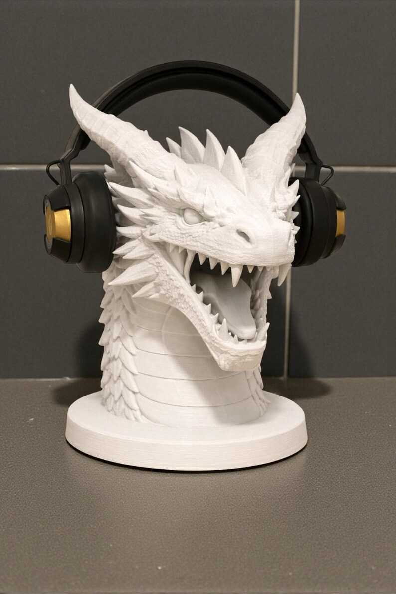 May include: A white dragon head sculpture with detailed scales and sharp teeth, designed to hold headphones. The dragon's head is mounted on a circular base, with black and gold headphones resting on its horns.