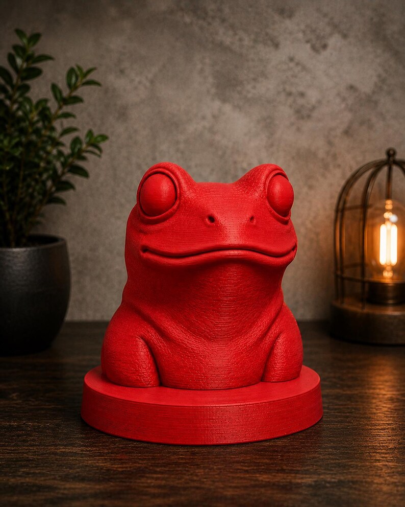 May include: A vibrant red frog sculpture with large, round eyes and a textured surface. The frog sits atop a matching red circular base. The sculpture is a decorative item, possibly for a desk or shelf.