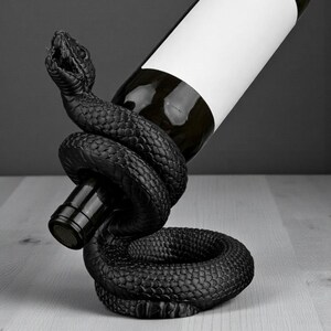 May include: A black snake wine bottle holder. The snake is coiled around a dark wine bottle with a blank white label. The holder is made of a textured material and sits on a light wood-grain surface. The background is a solid gray.