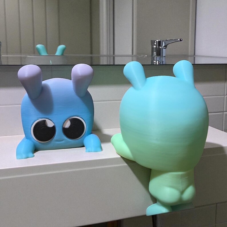 May include: Two colorful, cartoon-like figures with large eyes and rounded features. One is blue with purple ears, the other is a gradient of green and blue. They appear to be decorative items, possibly for a bathroom or children's room.