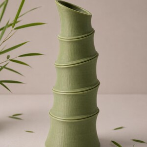 Puede incluir: Un jarr&oacute;n de cer&aacute;mica verde con forma de tallo de bamb&uacute;. El jarr&oacute;n tiene una superficie texturizada y una abertura curva. Hojas de bamb&uacute; dispersas alrededor del jarr&oacute;n.