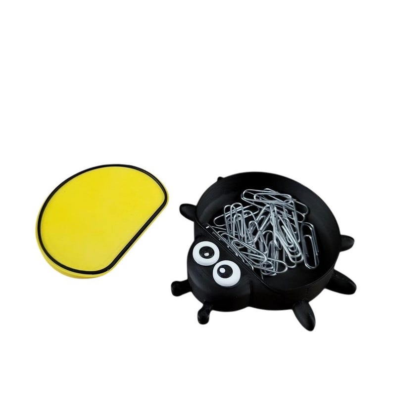 Puede incluir: Un organizador de escritorio negro con forma de mariquita lleno de clips plateados, junto a una alfombrilla de rat&oacute;n amarilla. La mariquita tiene ojos blancos y patas negras. La alfombrilla del rat&oacute;n es amarilla con un borde negro.