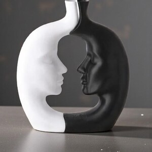 K&ouml;nnte beinhalten: Eine dekorative Vase in einem einzigartigen Design, das zwei sich gegen&uuml;berliegende menschliche Profile zeigt. Eine Seite ist wei&szlig;, die andere schwarz. Die Vase hat eine glatte Oberfl&auml;che und eine moderne &Auml;sthetik, geeignet f&uuml;r die Inneneinrichtung.